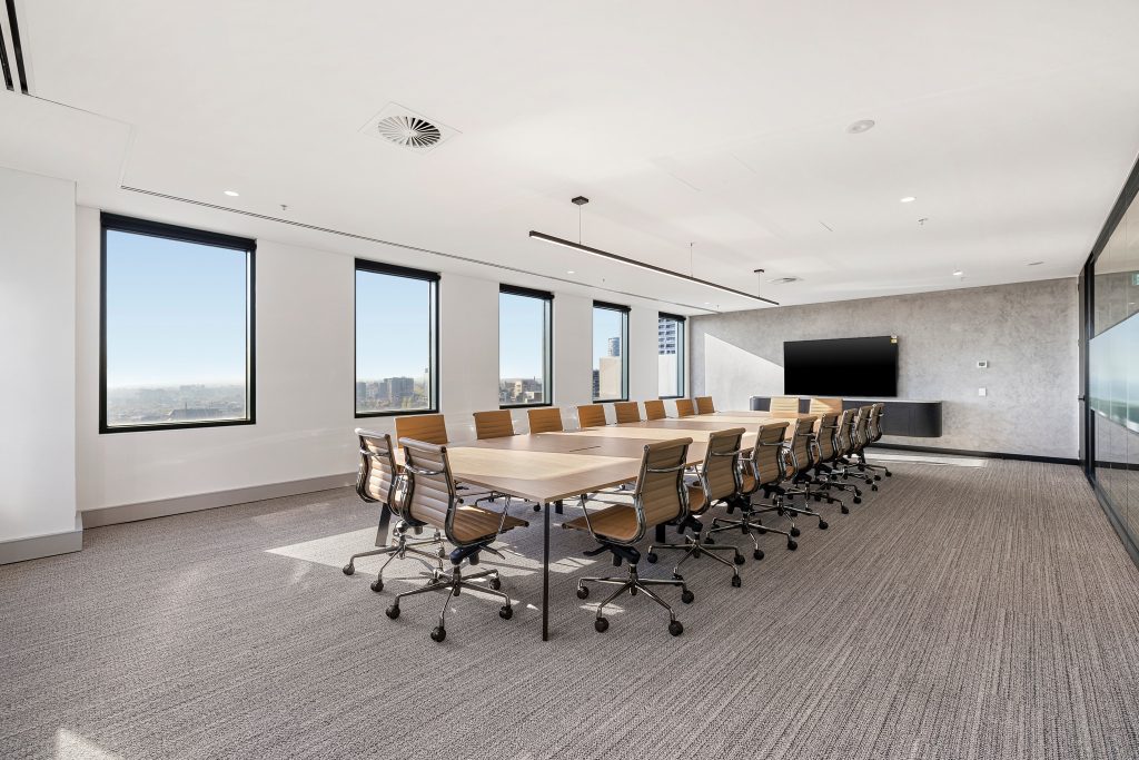A spacious conference room with a long wooden table surrounded by modern office chairs. The room features large windows providing a view of the cityscape, a wall-mounted TV screen, and sleek, minimalist decor. The floor is carpeted, and lighting is ample.