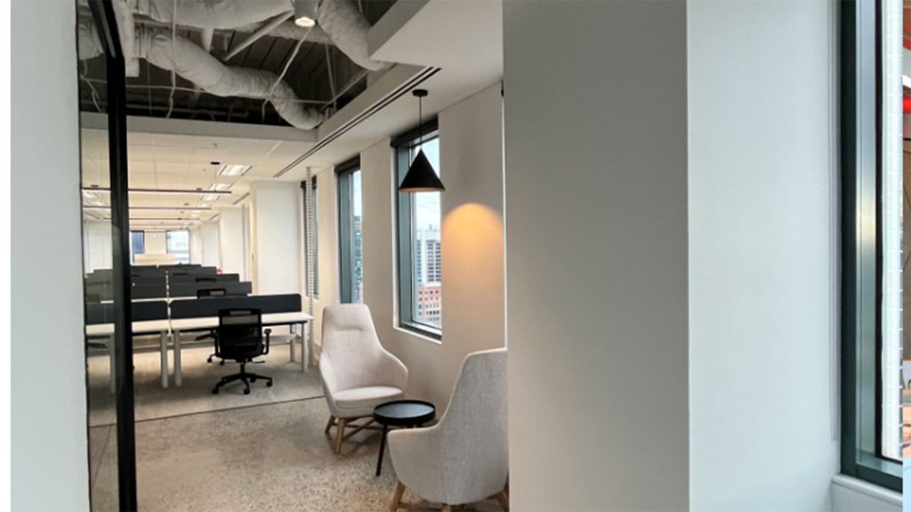 A modern office space with an open layout. In the foreground, there are two light grey chairs and a small round table near a large window, illuminated by a black hanging lamp. In the background, rows of desks with black office chairs can be seen.