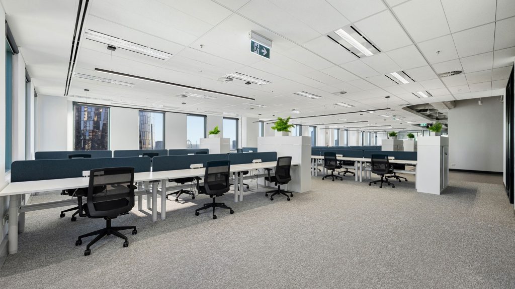 A spacious, modern office with rows of ergonomic chairs and long desks separated by partitions. The room is well-lit with large windows letting in natural light. Potted plants add greenery to the clean, minimalist design. The ceiling features recessed lighting.