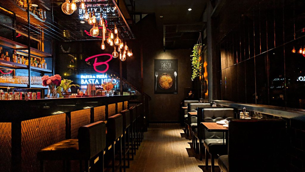 A cozy, dimly lit restaurant interior featuring a bar with stools on the left and cushioned booth seating on the right. Hanging Edison light bulbs provide a warm glow, and a neon sign reading "Basta Hiro" is visible above the bar. A framed artwork of pasta decorates the wall in the background.